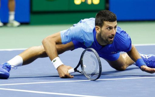 نوفاك دجوكوفيتش يؤدي حركة انزلاقية مذهلة أثناء إحدى مبارياته، مثبتًا جسده بالقرب من الأرض وممسكًا بمضربه، في لحظة تُظهر مرونته البدنية وتركيزه الذهني العالي.